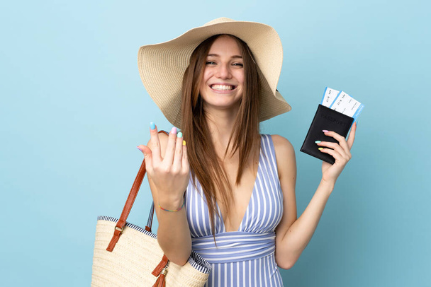 Młoda kaukaska kobieta w letnie wakacje trzymająca paszport odizolowany na niebieskim tle zapraszając do przybycia z ręką. Cieszę się, że przyszedłeś. - Zdjęcie, obraz