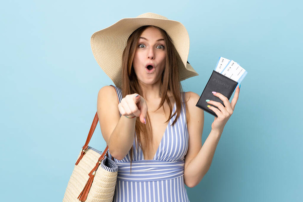 Junge kaukasische Frau in den Sommerferien mit Reisepass isoliert auf blauem Hintergrund überrascht und zeigt nach vorne - Foto, Bild