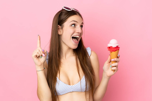 Молода біла жінка в купальнику тримає корнетне морозиво ізольовано на рожевому фоні, маючи намір реалізувати рішення піднімаючи палець вгору
 - Фото, зображення