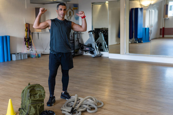 Motivated boot camp instructor stands with gym equipment in gym hall. Dumbbells, rope, sandbag on wooden floor. Instructor showing muscles. Portrait for gym concept. - Photo, Image