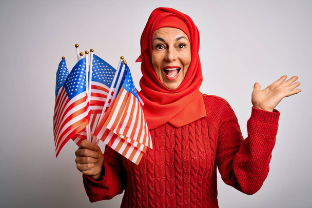 伝統的なイスラム教徒のヒジャーブを身に着けている中世の愛国的な女性は、米国の旗を保持し、非常に幸せと興奮,大きな笑顔で叫んで勝利を祝う勝者の表現と手を上げ - 写真・画像