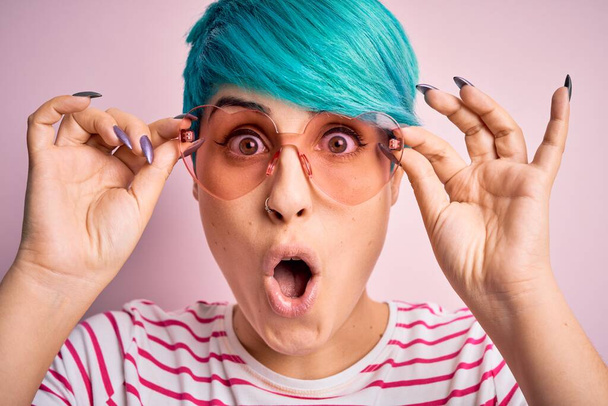 Young beautiful woman with blue fashion hair wearing fanny glasses with hearts scared in shock with a surprise face, afraid and excited with fear expression - Photo, Image