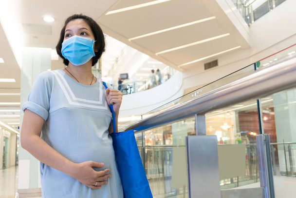 Pregnant Asian woman protect prevent infection herself from corona virus with wearing face mask and touching belly in the shopping mall. New normal life concept. - Foto, immagini