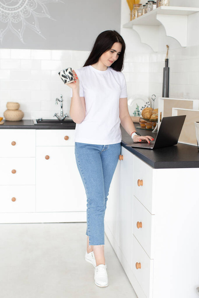 Egy fiatal nő laptopot használ és kávét iszik a konyhában reggel. Otthoni munka - Fotó, kép