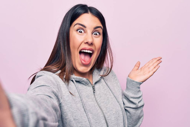 Young beautiful brunette sporty woman make selfie by the camera over pink background celebrating achievement with happy smile and winner expression with raised hand - Photo, Image