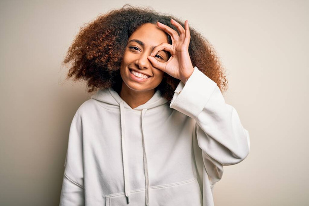 Junge afrikanisch-amerikanische Sportlerin mit Afro-Haaren trägt sportliches Sweatshirt und macht ok Geste mit lächelnder Hand, Augen blicken durch Finger mit glücklichem Gesicht. - Foto, Bild