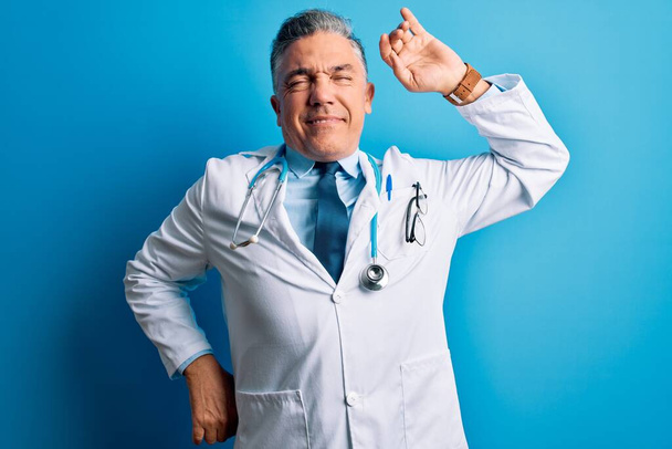 Middle age handsome grey-haired doctor man wearing coat and blue stethoscope stretching back, tired and relaxed, sleepy and yawning for early morning - Photo, Image