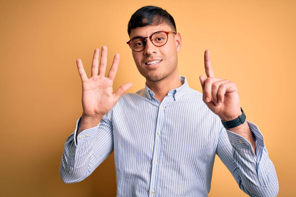 Jovem homem de negócios hispânico bonito usando óculos nerd sobre fundo amarelo mostrando e apontando para cima com os dedos número seis enquanto sorri confiante e feliz
. - Foto, Imagem