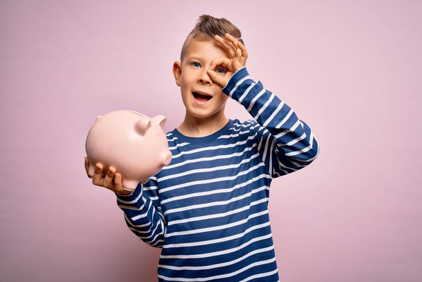 Молода маленька кавказька дитина бере гроші на скарбничку як заощадження багатства на рожевому тлі з щасливим обличчям, посміхаючись, роблячи добре знак з рукою на очах, дивлячись через пальці
 - Фото, зображення