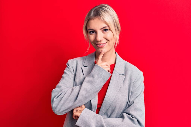 Jonge mooie blonde zakenvrouw draagt elegante jas over geïsoleerde rode achtergrond glimlachend zelfverzekerd kijkend naar de camera met gekruiste armen en hand op kin. Positief denken. - Foto, afbeelding