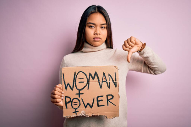 Giovane ragazza attivista asiatica che chiede diritti delle donne tenendo banner con il messaggio di potere della donna con la faccia arrabbiata, segno negativo che mostra antipatia con i pollici in giù, concetto di rifiuto
 - Foto, immagini