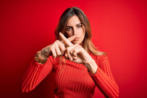 Mladá krásná brunetka žena na sobě ležérní rolák svetr přes červené pozadí Odmítnutí výraz křížení prsty dělá negativní znamení - Fotografie, Obrázek