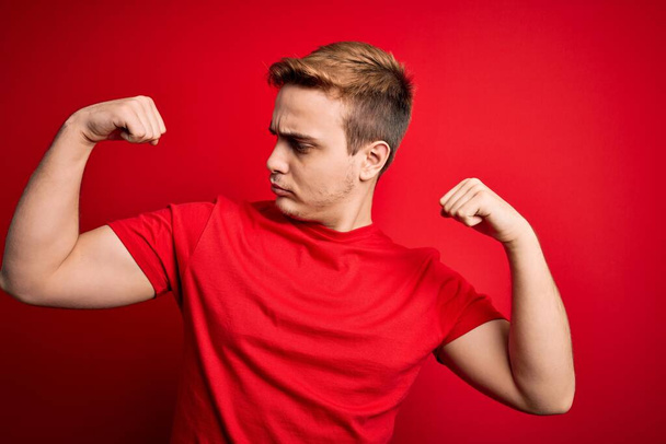 孤立した赤い背景の上にカジュアルなTシャツを着た若いハンサムな赤毛の男は腕の筋肉を誇りに思って笑顔を示しています。フィットネスコンセプト. - 写真・画像