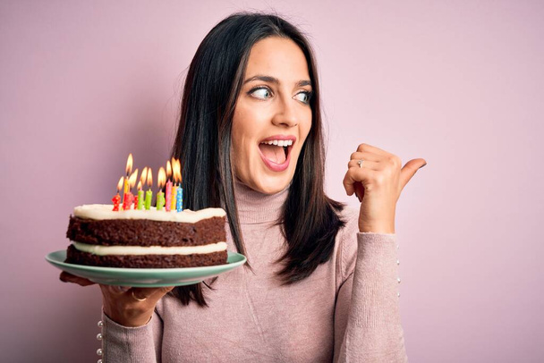 Junge Frau mit blauen Augen hält Geburtstagstorte mit Kerzen auf rosa Hintergrund und zeigt mit Daumen zur Seite mit glücklichem Gesicht lächelnd - Foto, Bild
