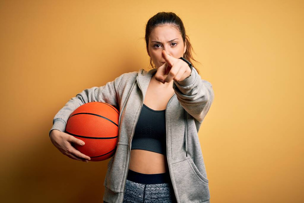 Молода красива брюнетка спортсменка тримає баскетбольний м'яч над жовтим тлом, вказуючи пальцем на камеру і до вас, знак руки, позитивний і впевнений жест спереду
 - Фото, зображення