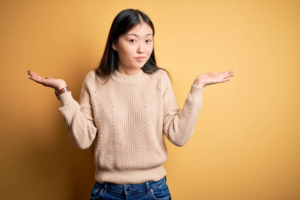 Young beautiful asian woman wearing casual sweater over yellow isolated background clueless and confused expression with arms and hands raised. Doubt concept. - Photo, Image