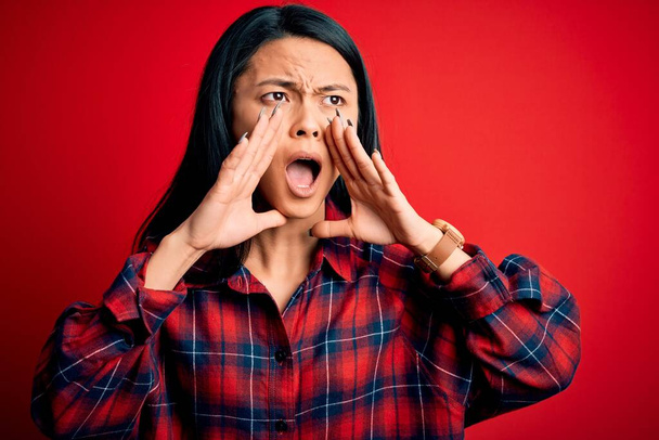 Молодая красивая китаянка в повседневной рубашке на изолированном красном фоне кричит сердито с руками над ртом
 - Фото, изображение