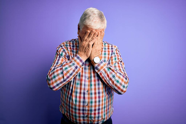 Starszy przystojny hoary mężczyzna ubrany w luźne kolorowe koszulki na odizolowanym fioletowym tle ze smutnym wyrazem twarzy pokrywającym ręce podczas płaczu. Koncepcja depresji. - Zdjęcie, obraz