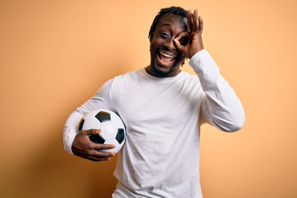 Fiatal afro-amerikai játékos férfi focizik kezében focilabda sárga háttér boldog arc mosolyog csinál rendben jel kéz a szemben néz keresztül ujjait - Fotó, kép
