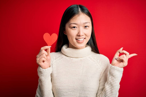 Joven mujer asiática sosteniendo romántico corazón rojo papel forma sobre rojo aislado fondo muy feliz señalando con la mano y el dedo a un lado
 - Foto, Imagen