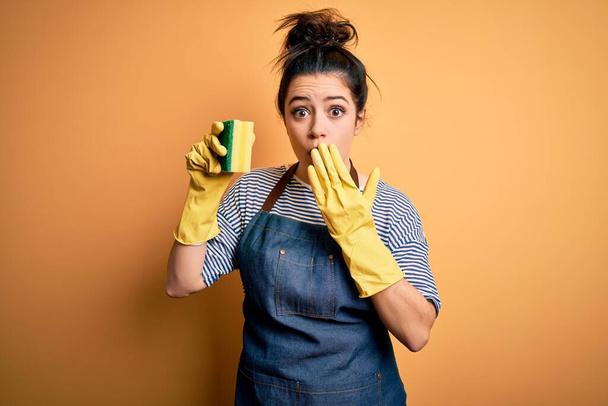 Mladá brunetka uklízečka žena nosí domácí rukavice drží scourer zakrýt ústa s rukou šokované hanbou za omyl, výraz strachu, strach v tichu, tajný koncept - Fotografie, Obrázek