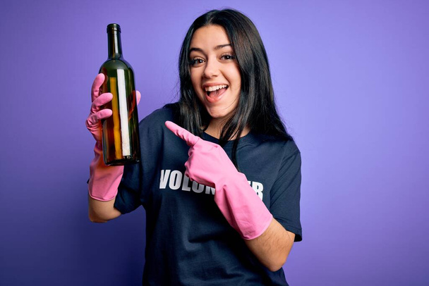 Giovane donna bruna indossa volontario t-shirt raccogliendo bottiglia di vetro su sfondo viola molto felice puntando con mano e dito
 - Foto, immagini