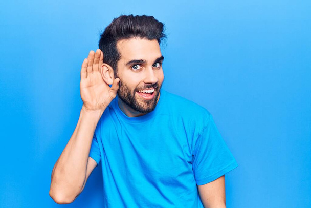Jeune homme beau à la barbe portant un t-shirt décontracté souriant avec la main sur l'oreille écoutant et entendant des rumeurs ou des commérages. concept de surdité.  - Photo, image