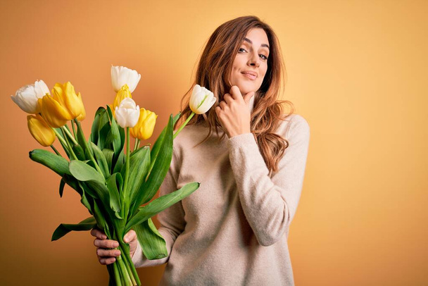 若い美しいブルネットの女性は、孤立した背景の上に黄色のチューリップの花束を持っており、カメラが交差した腕と手で笑みを浮かべていることに自信を持っている。肯定的な考え. - 写真・画像