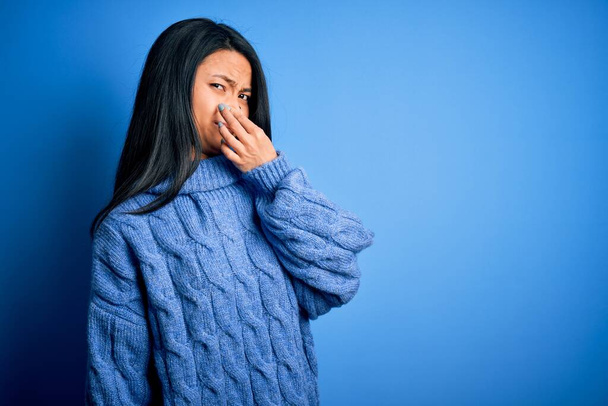 Joven mujer china hermosa usando suéter casual sobre fondo azul aislado oliendo algo apestoso y repugnante, olor intolerable, conteniendo la respiración con los dedos en la nariz. Mal olor
 - Foto, Imagen