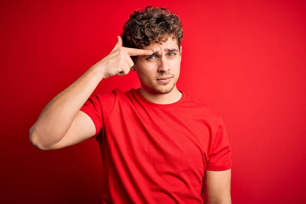 Young blond handsome man with curly hair wearing casual t-shirt over red background pointing unhappy to pimple on forehead, ugly infection of blackhead. Acne and skin problem - Photo, Image