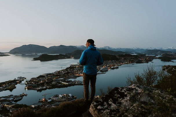 Hiker is observing picturesque norwegian city scenery from the mountain. Travel and hiking concept. Shot during the blue hour. - Фото, изображение