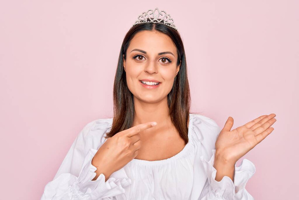 Young beautiful brunette woman wearing princess tiara standing over isolated pink background amazed and smiling to the camera while presenting with hand and pointing with finger. - Photo, Image
