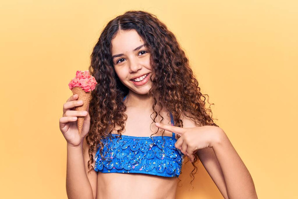 Belle fille enfant aux cheveux bouclés portant un bikini tenant la crème glacée pointant du doigt vers un sourire heureux et fier  - Photo, image