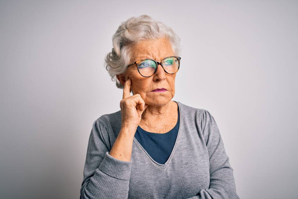 Senior kaunis harmaa tukka nainen yllään rento villapaita ja lasit valkoisella taustalla käsi leuka ajatella kysymys, mietteliäs ilme. Hymyillen ajattelevaisilla kasvoilla. Epävarma käsite. - Valokuva, kuva