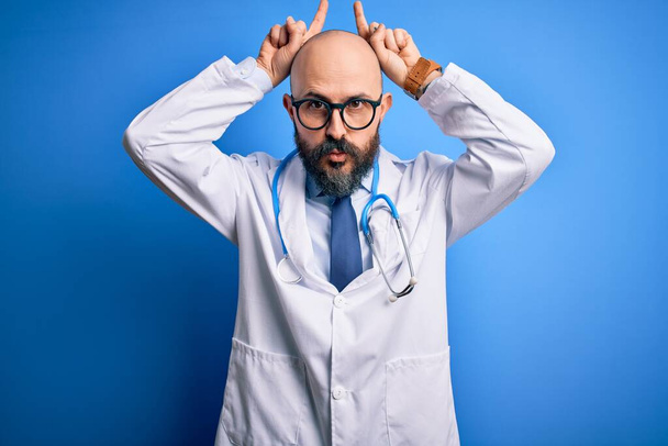 Komea kalju lääkäri, jolla on parta silmälasit ja stetoskooppi sinisen taustan päällä, tekee hauskoja eleitä sormella pään päällä kuin härän sarvet. - Valokuva, kuva