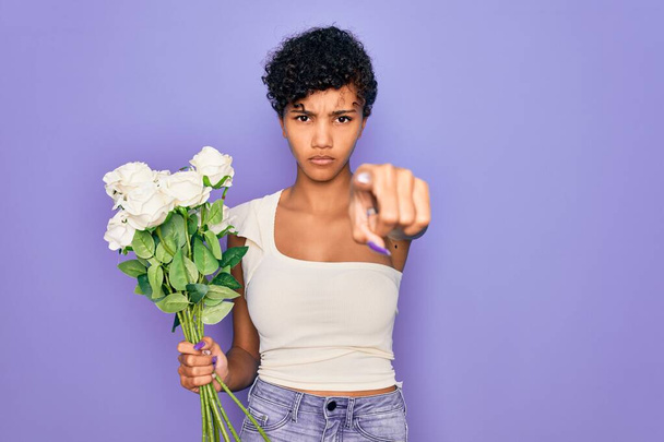 Beautiful african american afro woman holding bouquet of white flowers over purple background pointing with finger to the camera and to you, hand sign, positive and confident gesture from the front - Фото, изображение