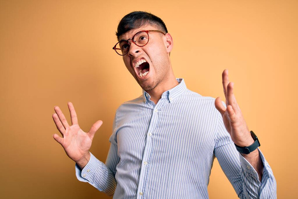 Jovem homem de negócios hispânico bonito usando óculos nerd sobre fundo amarelo louco e louco gritando e gritando com expressão agressiva e braços levantados. Conceito de frustração
. - Foto, Imagem