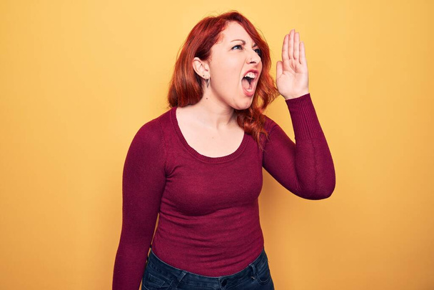 若い美しい赤毛の女性は、黄色の背景の叫びに立ってカジュアルなセーターを着て、口の上に手で横に大声で叫んでいます。コミュニケーションの概念. - 写真・画像