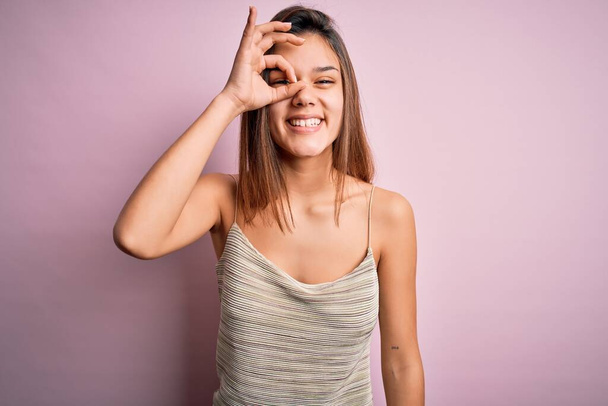 Jeune belle fille brune portant un t-shirt rayé décontracté sur fond rose isolé faisant un geste correct avec le sourire de la main, les yeux regardant à travers les doigts avec un visage heureux
. - Photo, image