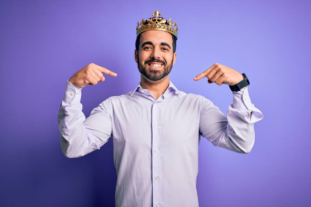 Jonge knappe man met baard dragen gouden kroon van koning over paarse achtergrond kijken zelfverzekerd met glimlach op het gezicht, wijzend zichzelf met vingers trots en gelukkig. - Foto, afbeelding