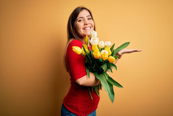 Mladá blondýnka drží romantickou kytici tulipánů květy přes žluté pozadí ukazuje stranou s otevřenými dlaněmi ukazující kopírovací prostor, prezentující reklamu s nadšeným úsměvem - Fotografie, Obrázek
