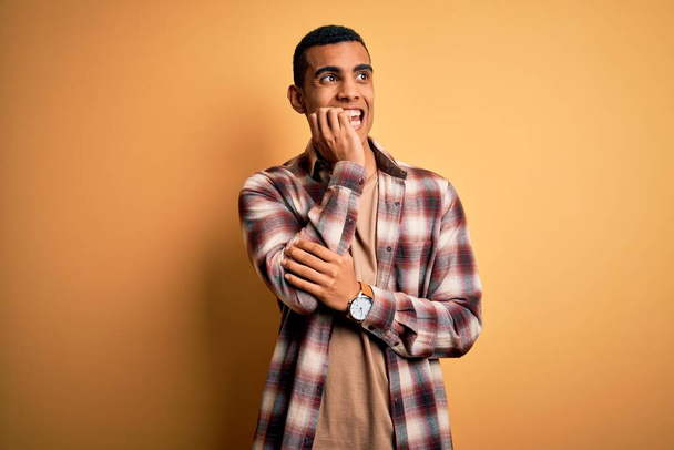 Young handsome african american man wearing casual shirt standing over yellow background looking stressed and nervous with hands on mouth biting nails. Anxiety problem. - Foto, immagini