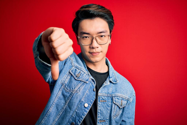 赤い背景にデニムジャケットと眼鏡をかけている若いハンサムな中国人男性は、親指を下にジェスチャーで不満と否定を示して怒っているように見えます。悪い表現. - 写真・画像