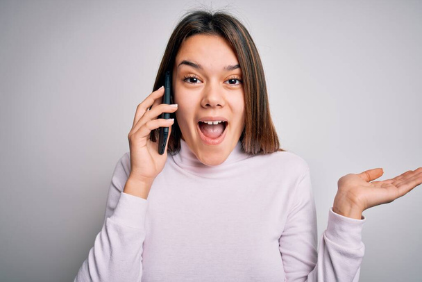 Beautiful brunette girl having conversation talking on the smartphone over white background very happy and excited, winner expression celebrating victory screaming with big smile and raised hands - Photo, image