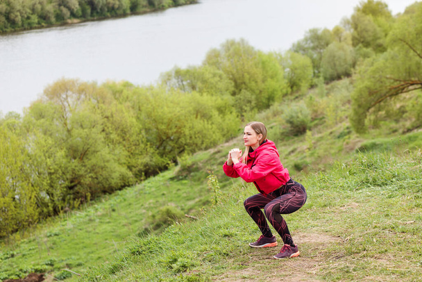 Athletic caucasian woman doing squats on the river bank. Sporty lifestyle - Photo, Image