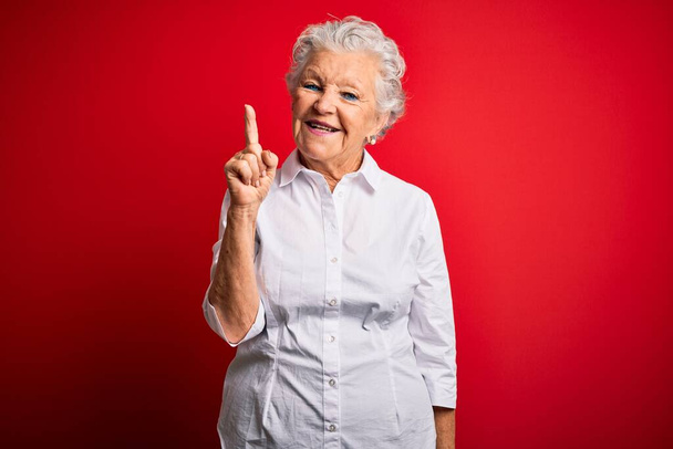 Старшая красивая женщина в элегантной рубашке стоит на изолированном красном фоне показывая пальцем вверх с успешной идеей. Взволнован и счастлив. Номер один
. - Фото, изображение