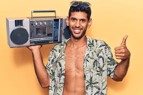 Fiatal latin férfi visel nyári ing gazdaság boombox mosolygós boldog és pozitív, hüvelykujj fel csinál kiváló és jóváhagyási jel  - Fotó, kép
