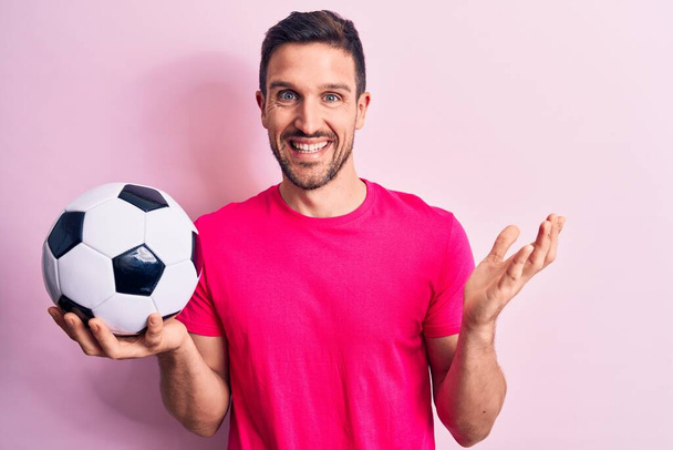 Young handsome player man playing soccer holding football ball over isolated pink background celebrating achievement with happy smile and winner expression with raised hand - Фото, изображение