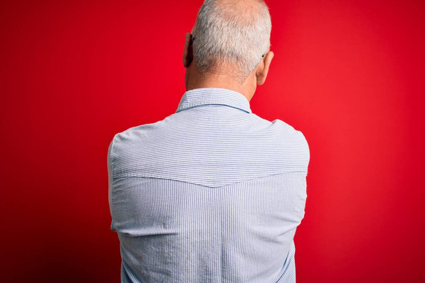 Hombre guapo hoary de mediana edad con camisa a rayas casual y gafas sobre fondo rojo de pie hacia atrás mirando hacia otro lado con los brazos cruzados
 - Foto, imagen