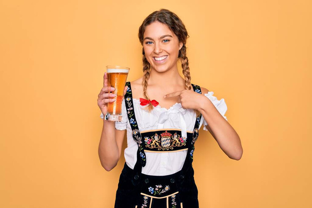 Mavi gözlü, güzel sarışın Alman kadın, Ekim Festivali elbisesi giyiyor. Bir bardak bira içiyor ve kendini işaret ediyor. - Fotoğraf, Görsel